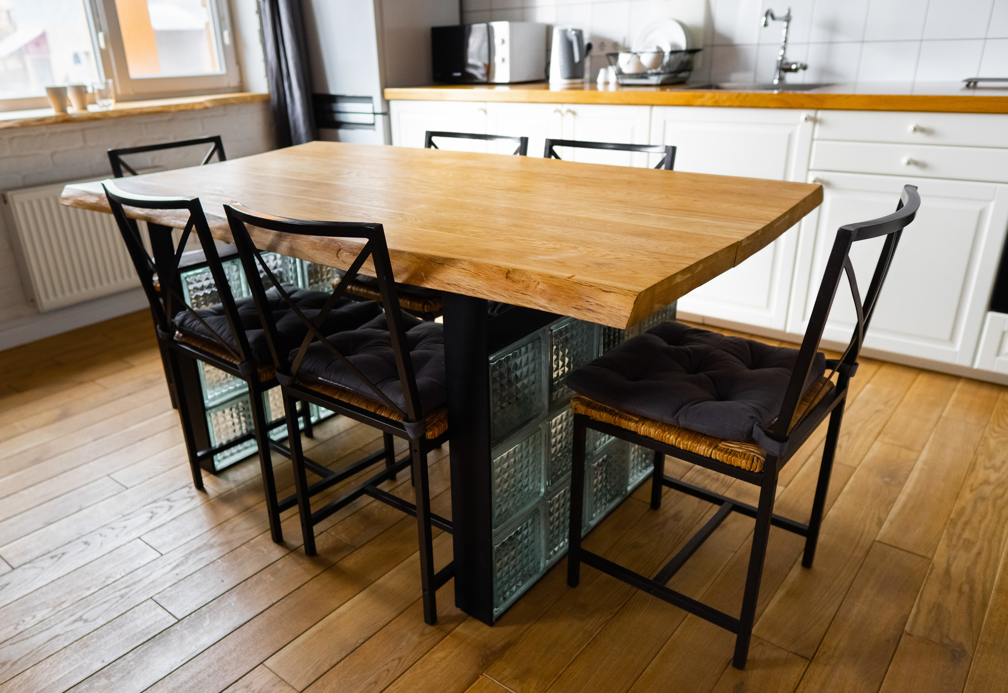 A big wooden dining table with glass blocks and metal wicker chairs and pillows in modern scandinavian an eat-in kitchen, against bright white furnitures, appliances and light wood floor